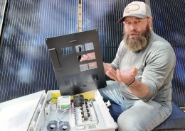 Bearded man in a gray shirt kneels beside an open solar power inverter, showing wiring, with tall solar panels in the background.
