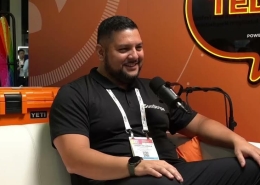 Man with short hair and beard sits on a white couch, speaking into a microphone during an interview setup at an event.