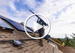 Two workers install solar panels on a sloped residential roof under a blue sky with clouds.