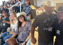 Crowd at a country concert: people wearing cowboy hats in stadium seating, smiling and holding drinks in the foreground. Includes two women in hats and sunglasses.